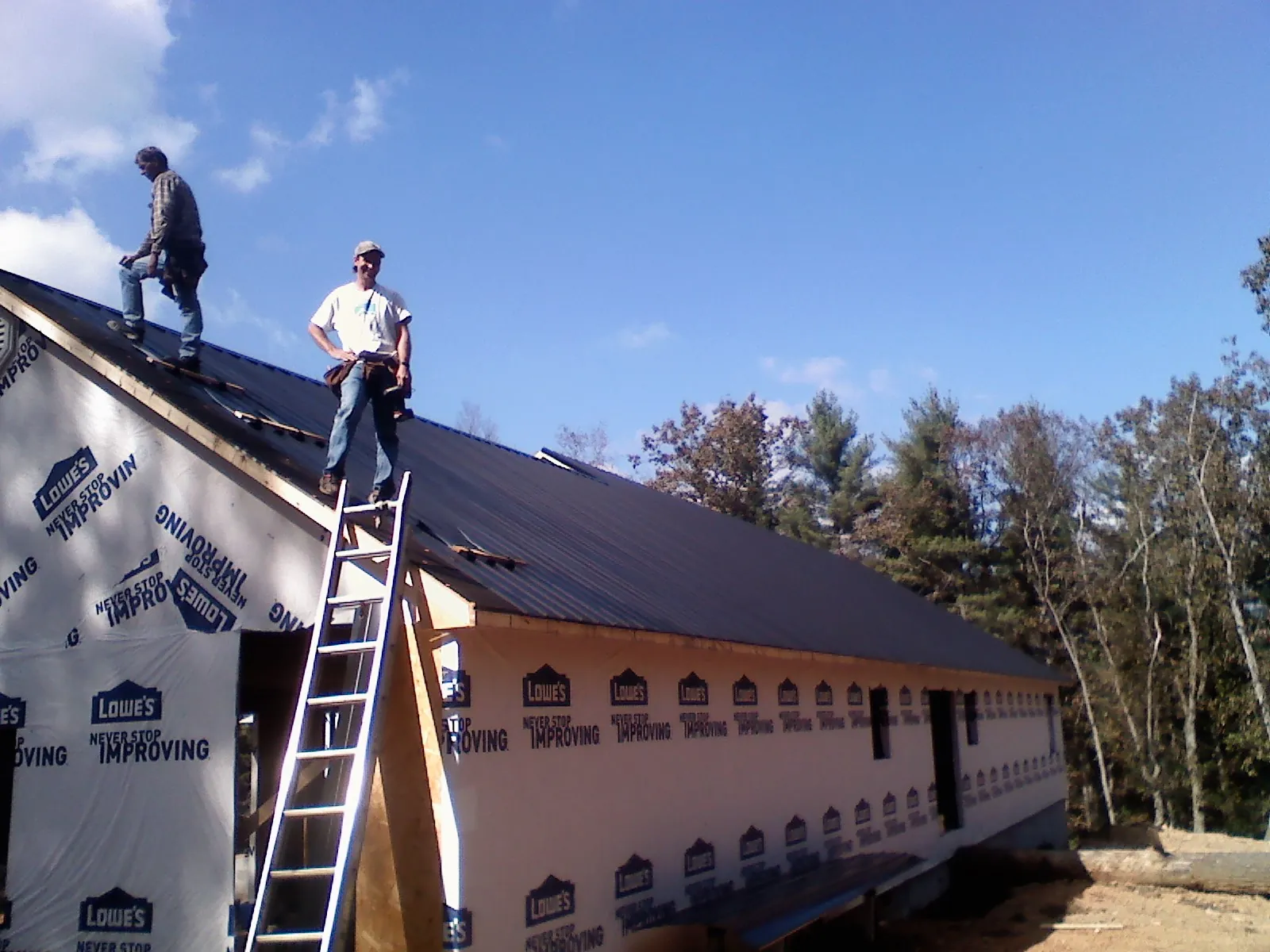 Two roofers standing on a long dark metal roof over a wrapped building in Badin